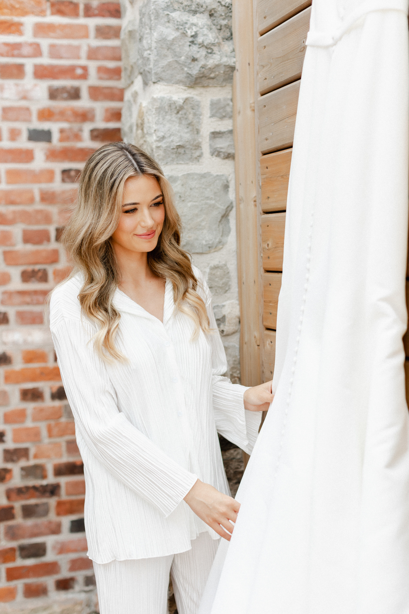 A bride in white pajamas gazes at her wedding dress.