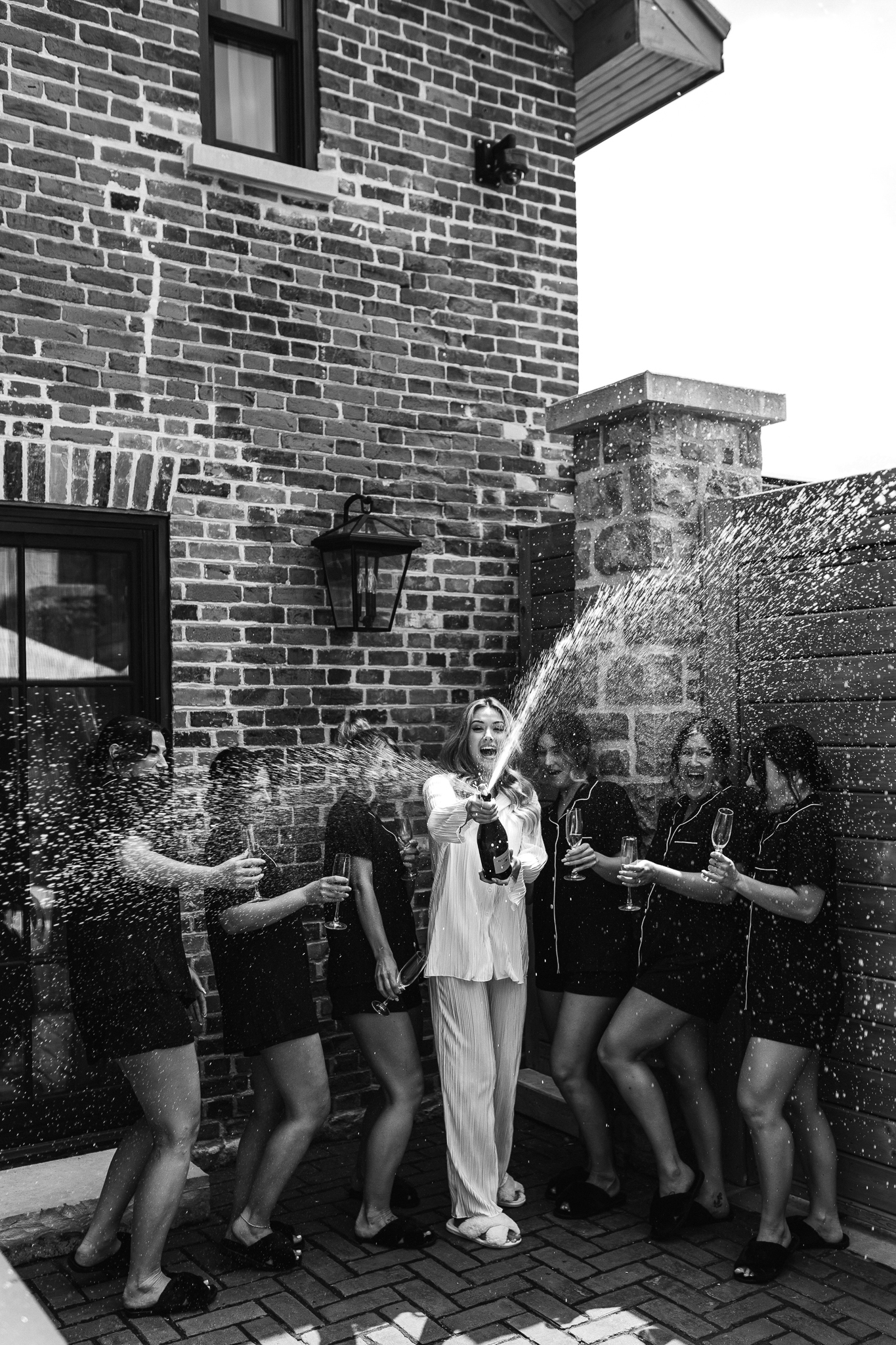 Bridesmaids and Bride pose in front of a brick building, while they pop a bottle of champagne.