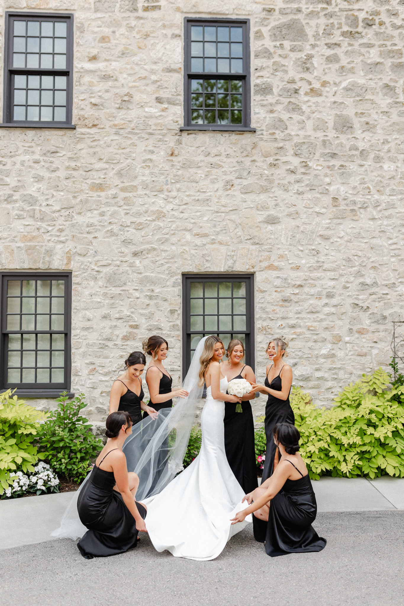 Bride and bridesmaids smile for a portrait outside a stone house, showcasing the brides dress