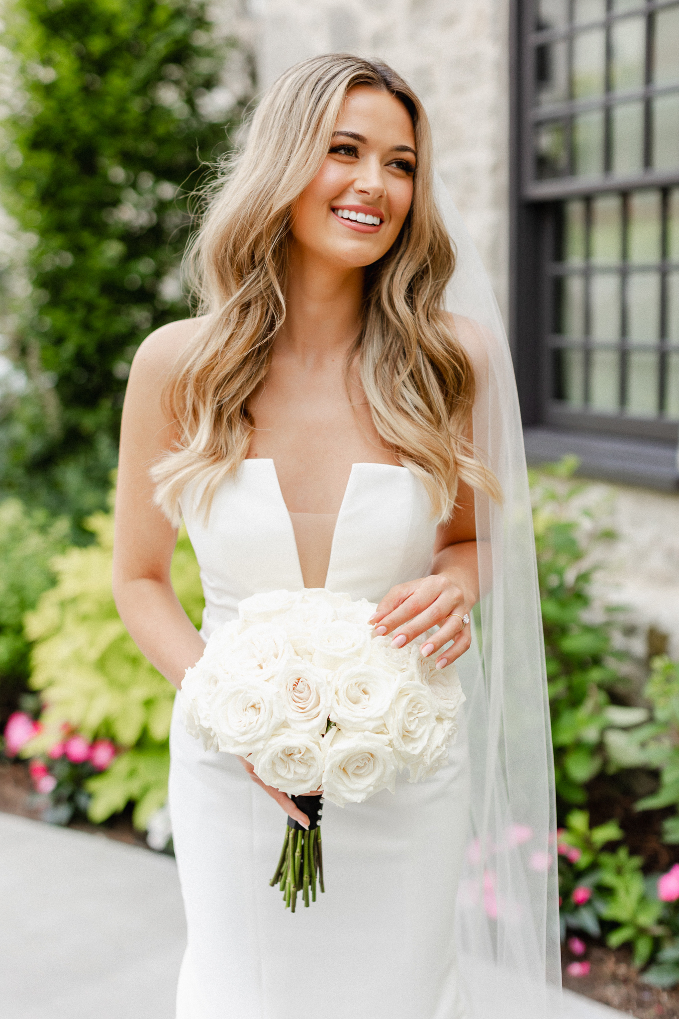 A stunning bride, elegantly dressed in white, gracefully holds a bouquet and touches it, showcasing her wedding ring while laughing