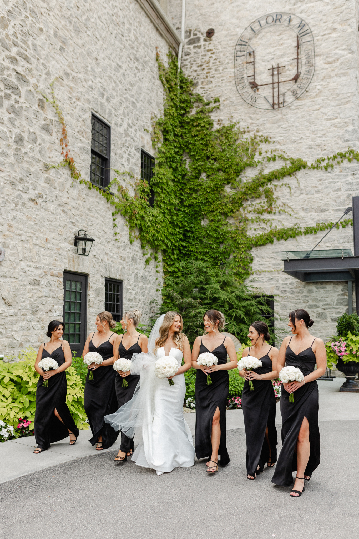 Bridesmaids elegantly dressed in black attire, holding beautiful white bouquets with the bride while walking and looking at each other