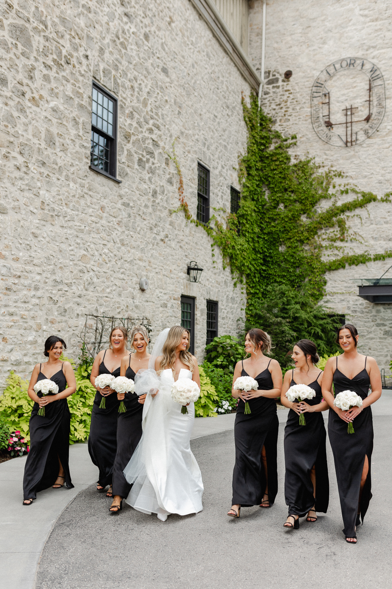 Bridesmaids elegantly dressed in black attire, holding beautiful white bouquets with the bride while walking and looking at each other