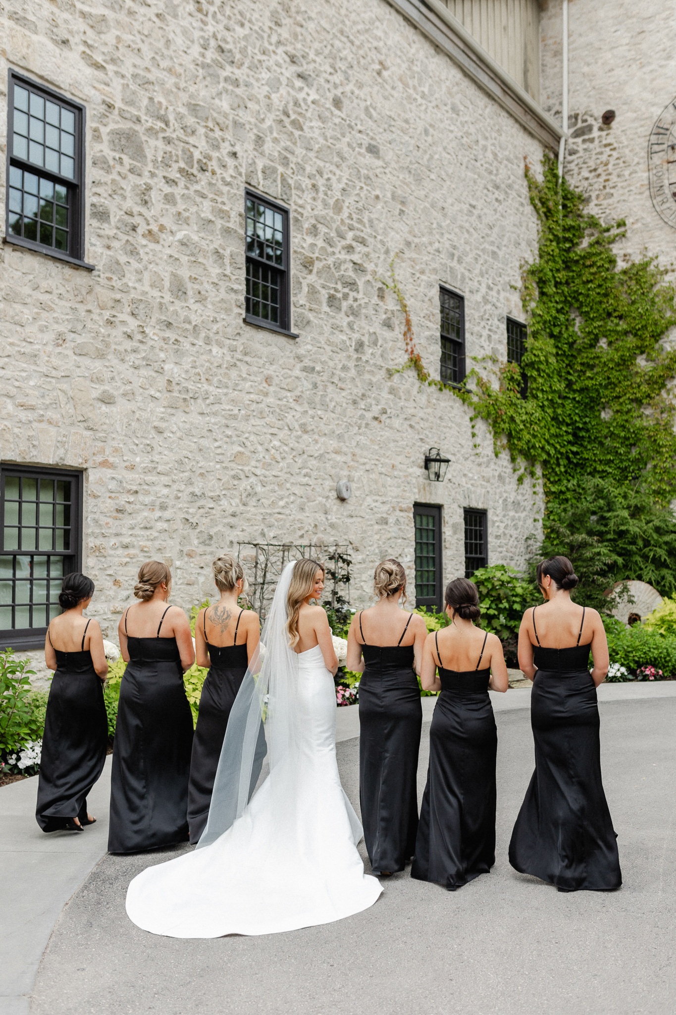 Bridesmaids elegantly dressed in black attire, holding beautiful white bouquets with the bride while walking away from the camera