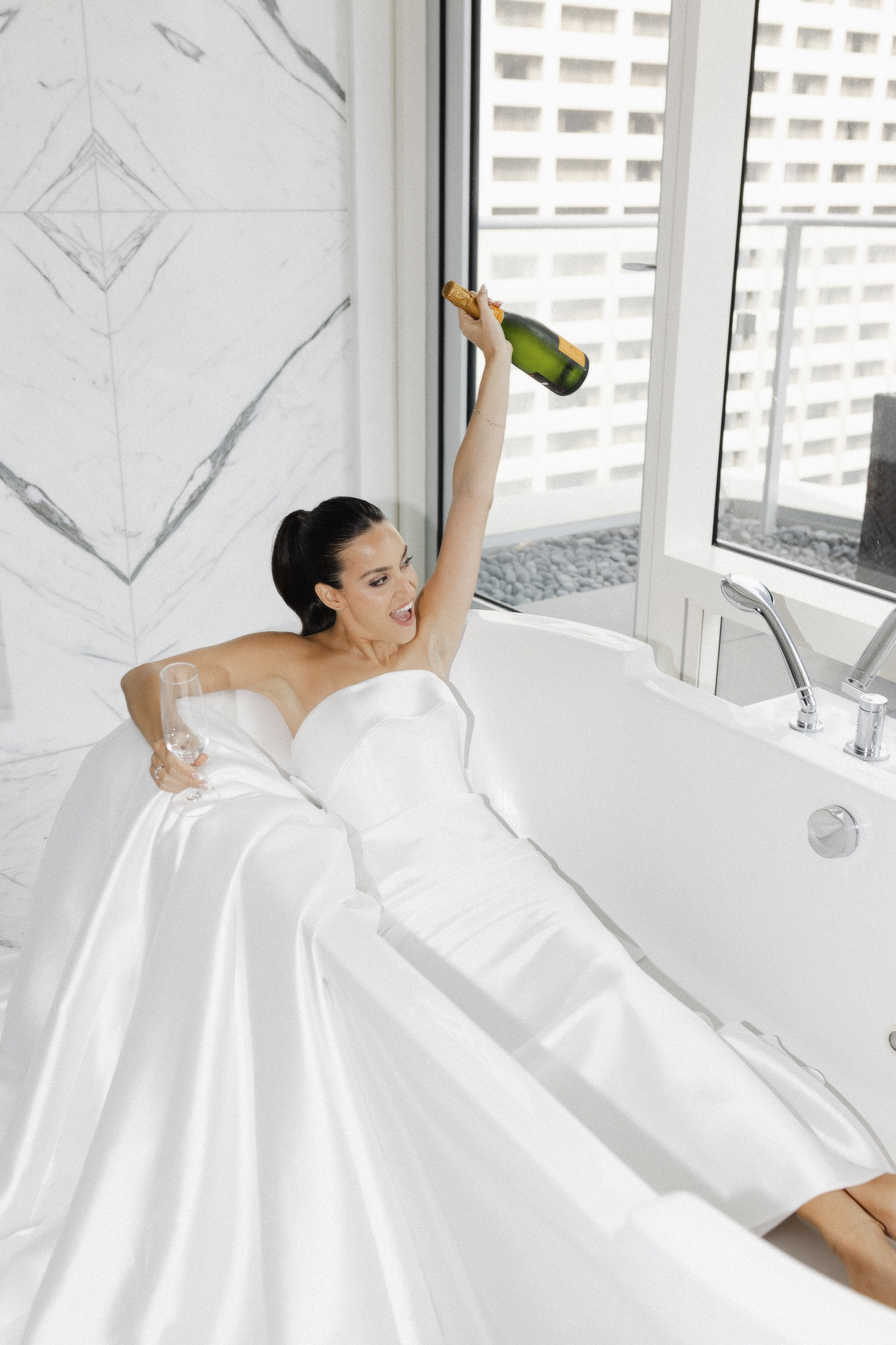 The bride in a wedding dress holding a bottle of champagne in a white bathtub.