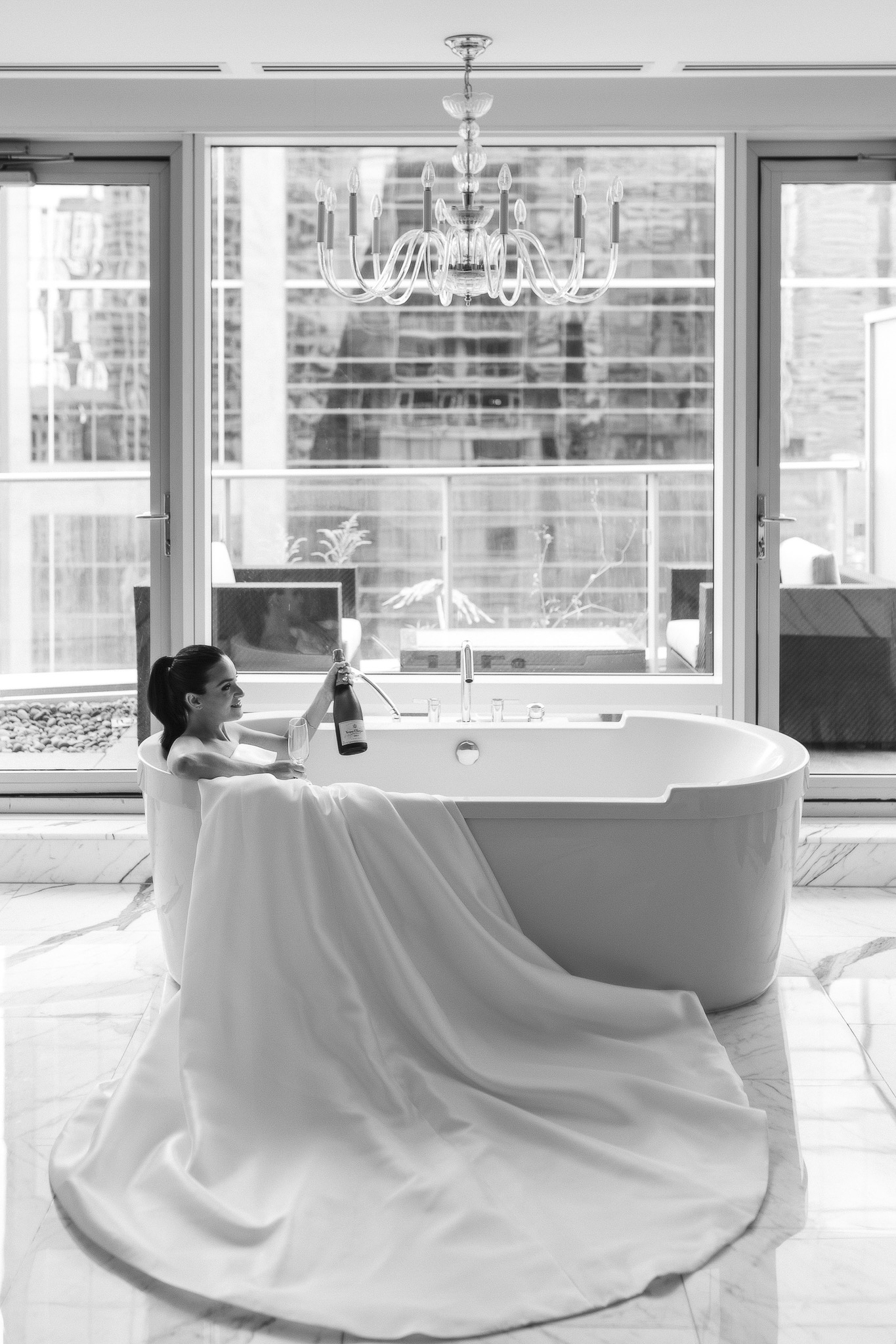 The bride in a wedding dress holding a bottle of champagne in a white bathtub, the dress hanging over the side of the tub.