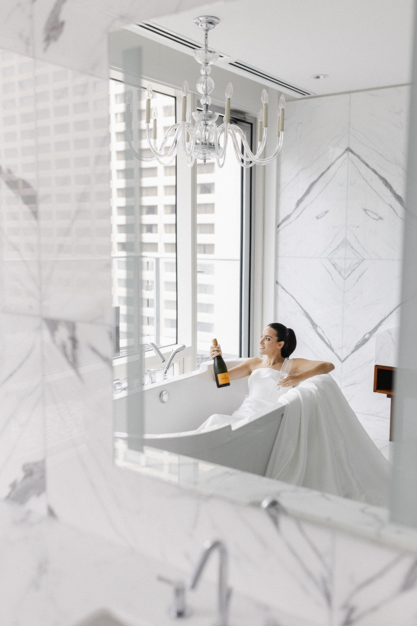 The bride in a wedding dress holding a bottle of champagne in a white bathtub, shot through a mirror reflection.