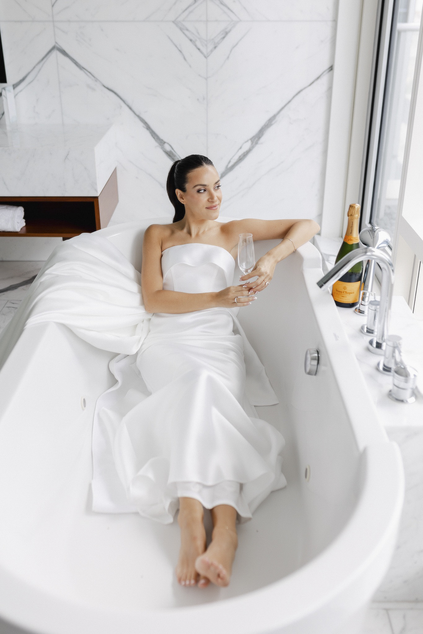 The bride in a wedding dress holding a champagne glass in a white bathtub
