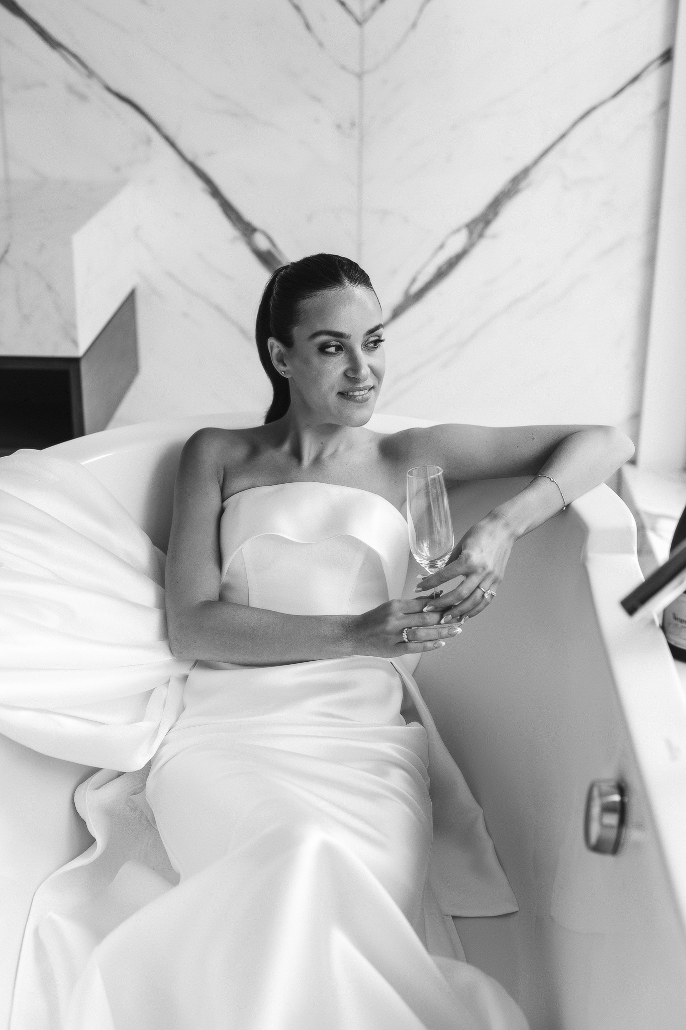 The bride in a wedding dress holding a champagne glass in a white bathtub