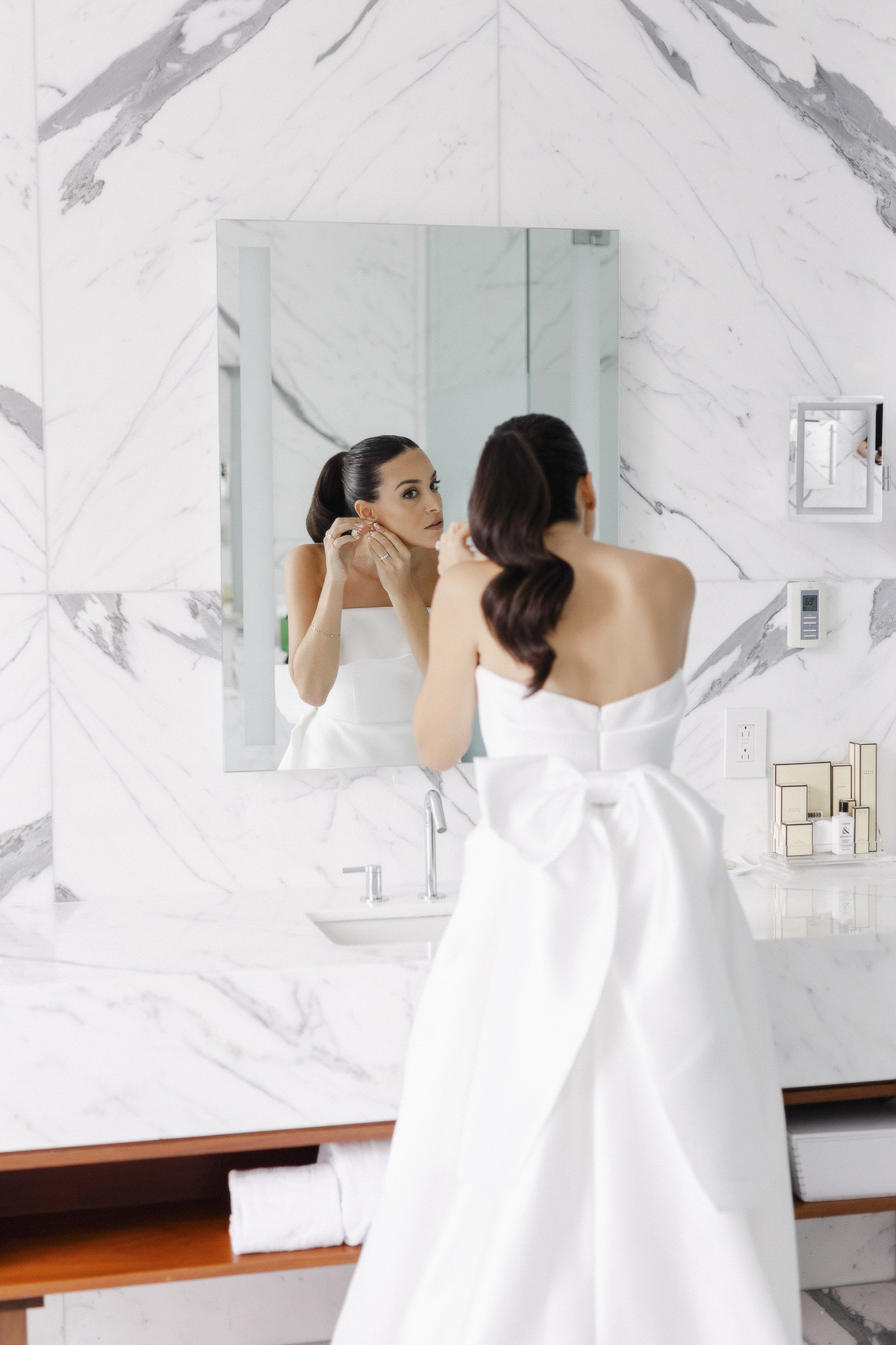 Bride putting on earrings in a bathroom mirror.