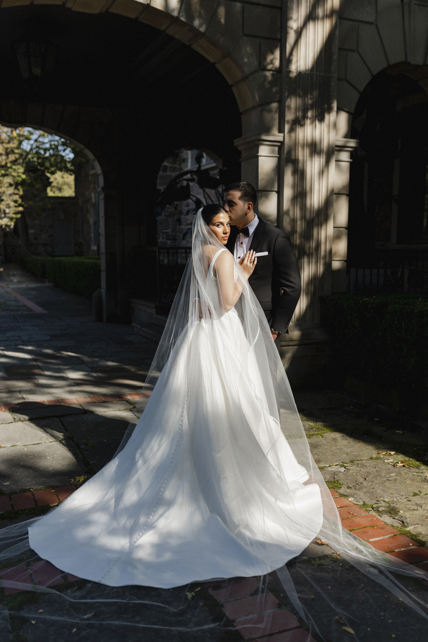 Bride and groom standing under a stone archway in Graydon Hall Manor with the bride’s veil flowing behind her, captured in a dramatic, editorial wedding photography style