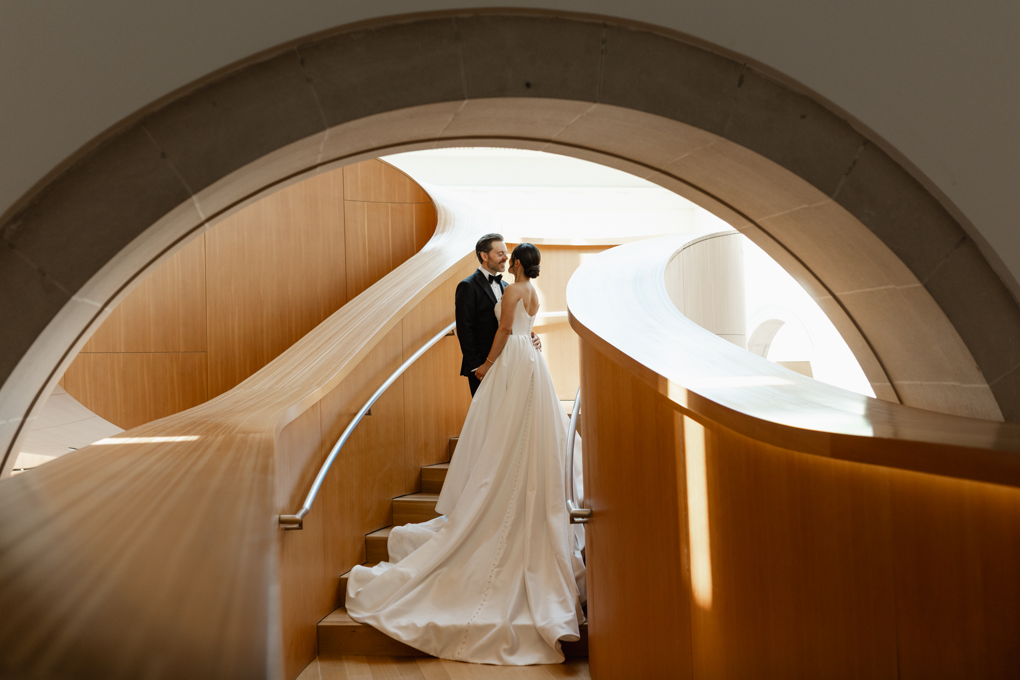 Bride and groom standing together on a curved wooden staircase in a modern architectural interior of AGO museum, captured in an editorial, elegant wedding portrait style