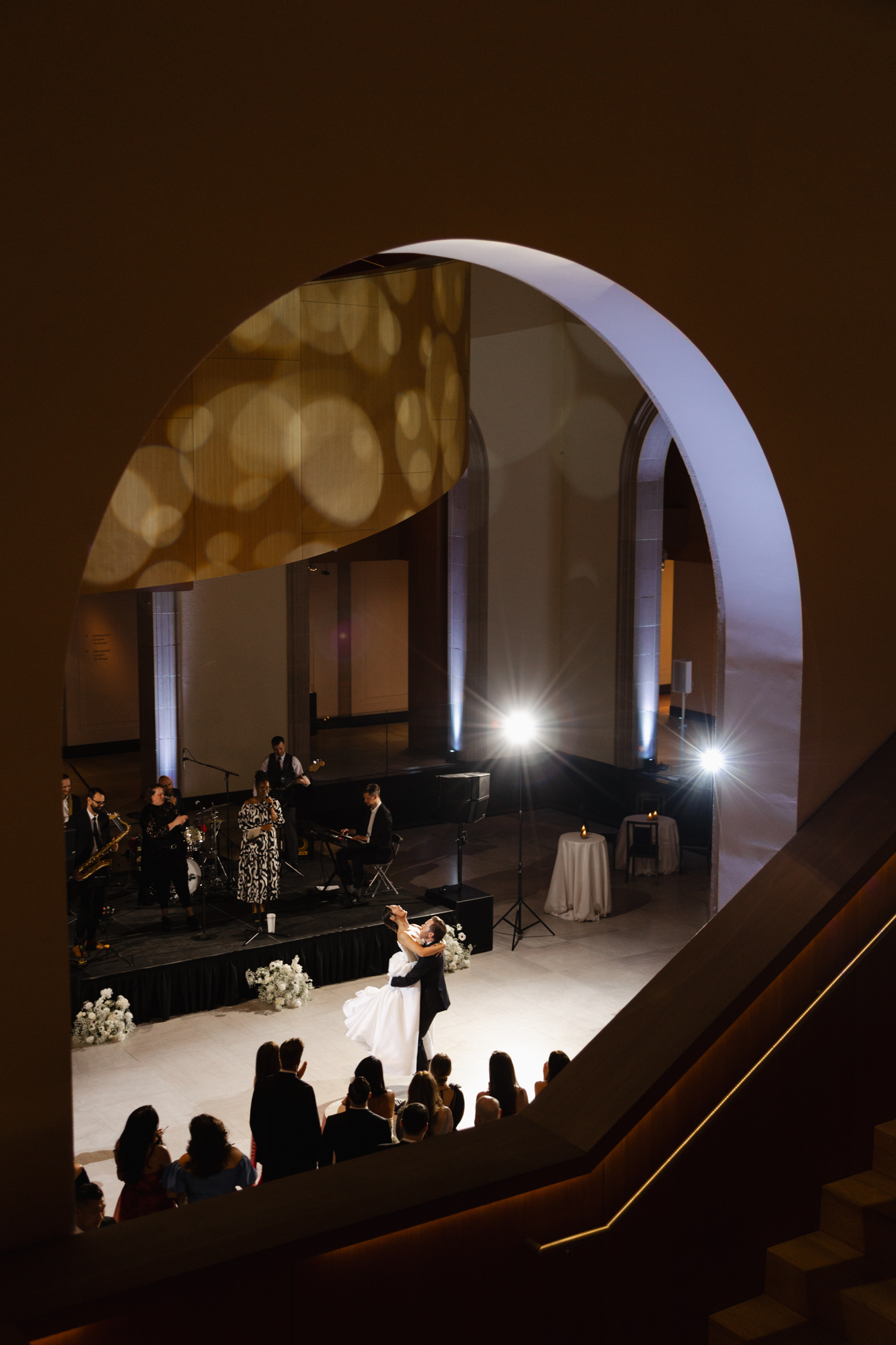 Bride and groom sharing their first dance on a stage in AGO with live musicians, viewed through an architectural arch, captured in a cinematic, editorial wedding style