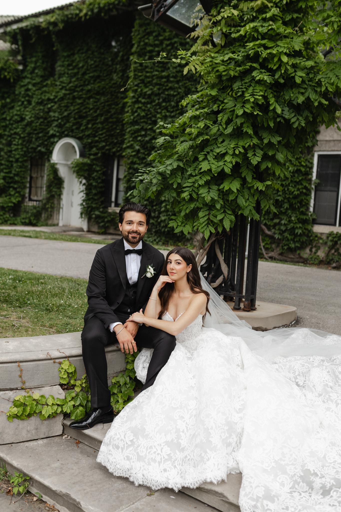 Bride and groom seated together outside an ivy-covered historic building, captured in a composed, editorial wedding portrait style with soft natural light.