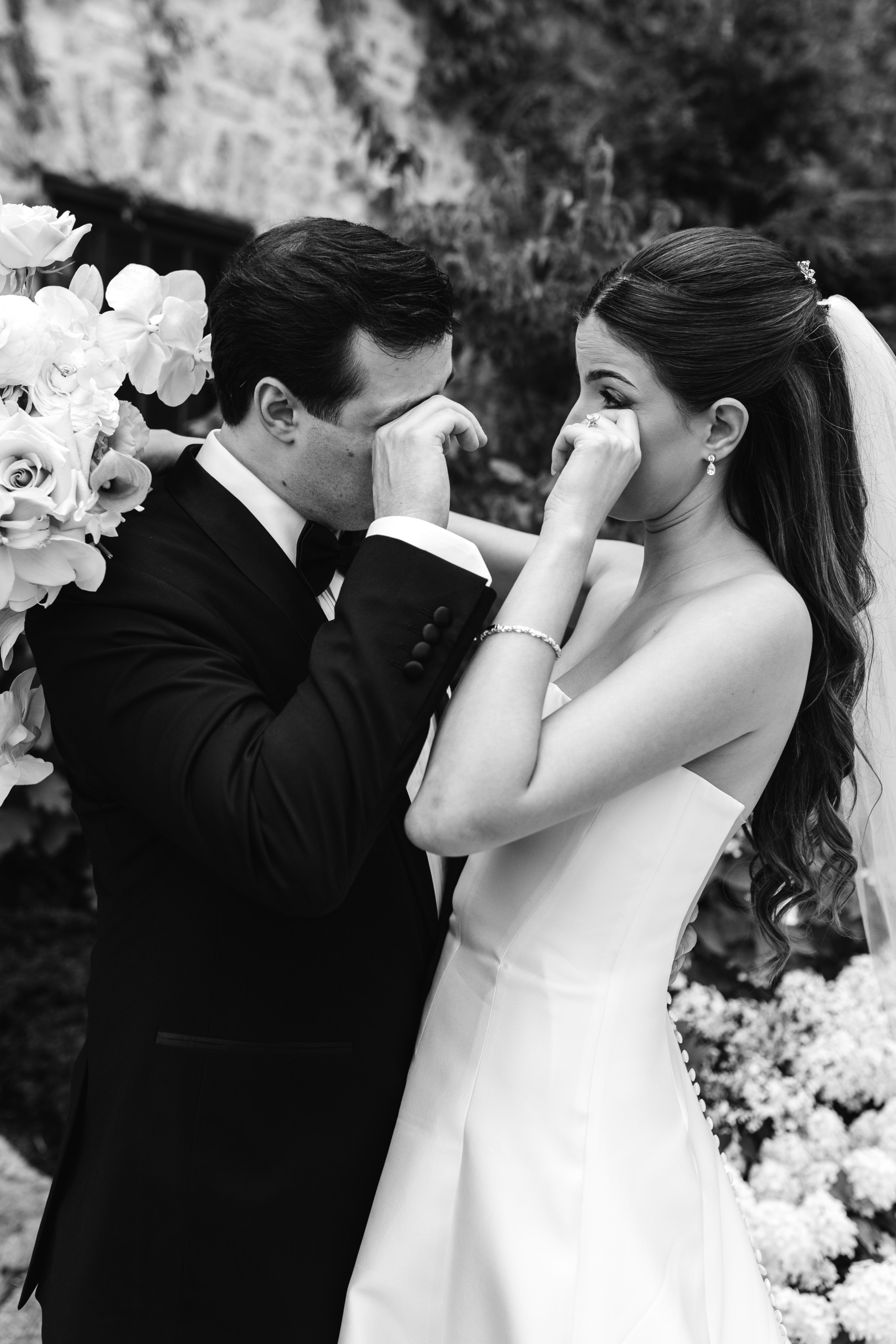 Candid black and white portrait of a bride and groom wiping away tears while laughing together, captured in an intimate, unposed wedding moment.
