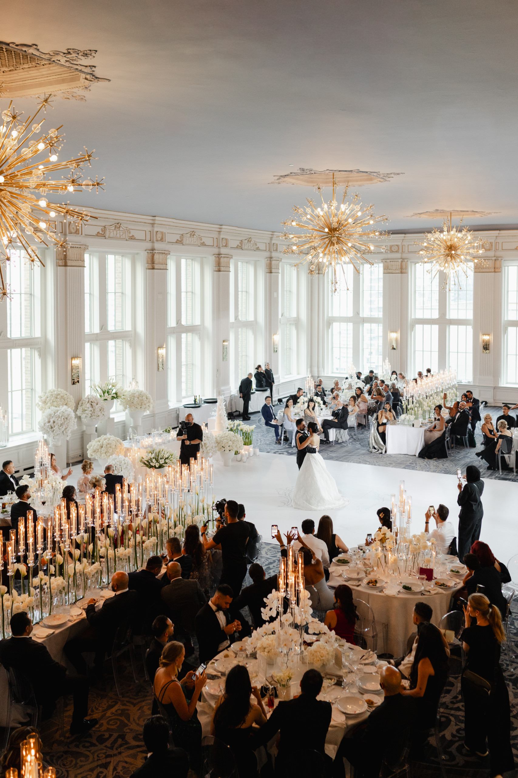 Wide photojournalistic view of a wedding reception in a grand ballroom with guests seated, tall floral centerpieces, candlelight, and a couple sharing a moment at the center of the room under chandelier light