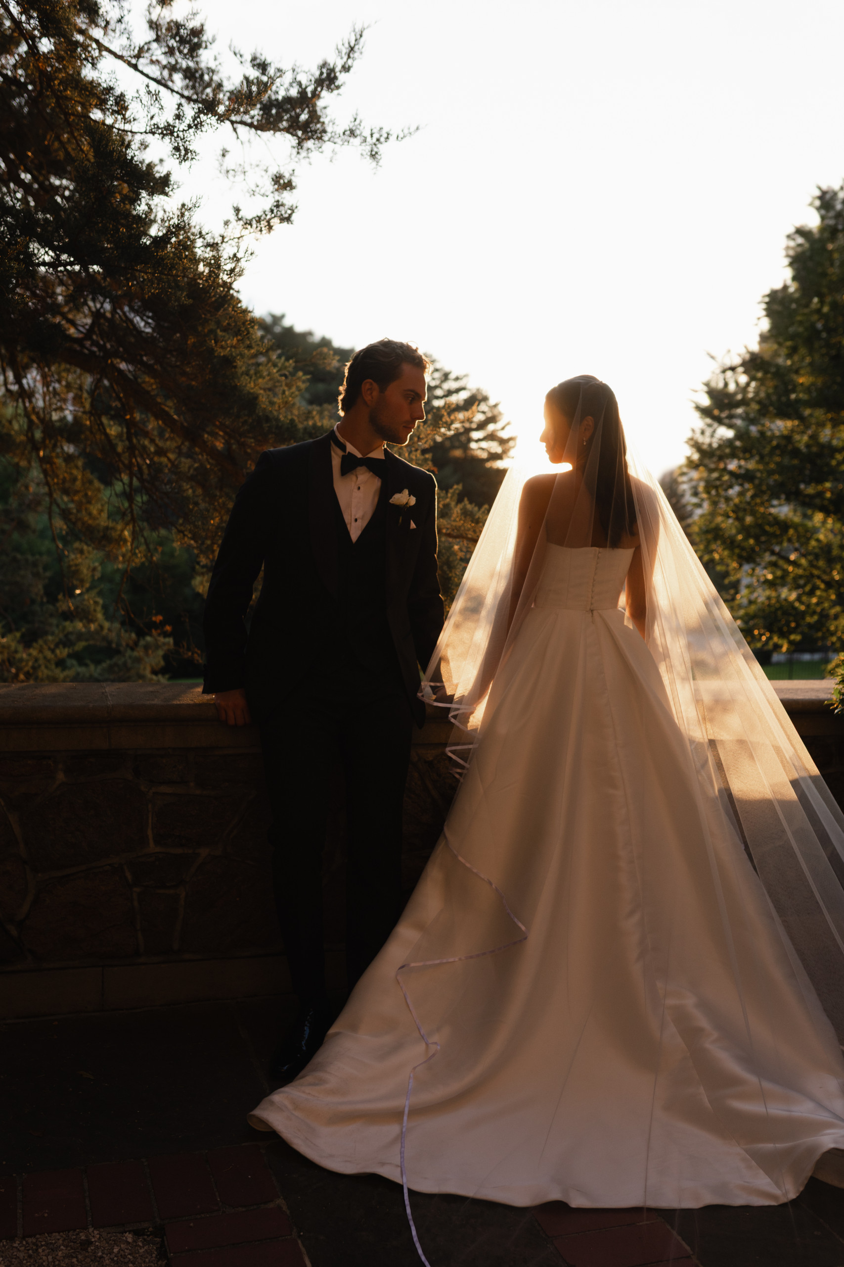 Bride and groom standing together at sunset with the bride’s veil glowing in backlight, captured in a refined, editorial wedding photography style.