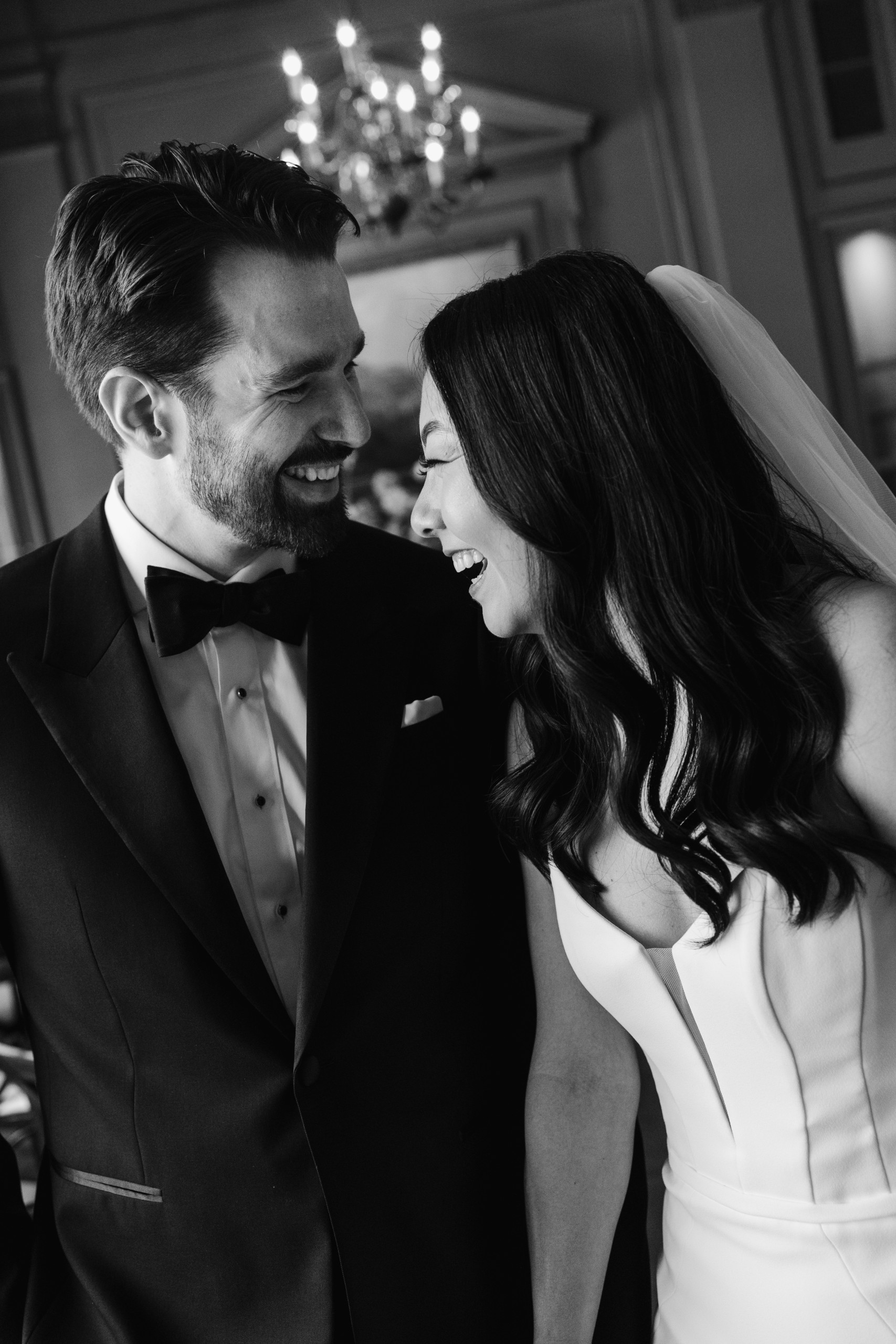 Bride and groom laughing together during an intimate indoor wedding moment, captured in black and white with soft chandelier light in an editorial, photojournalistic style