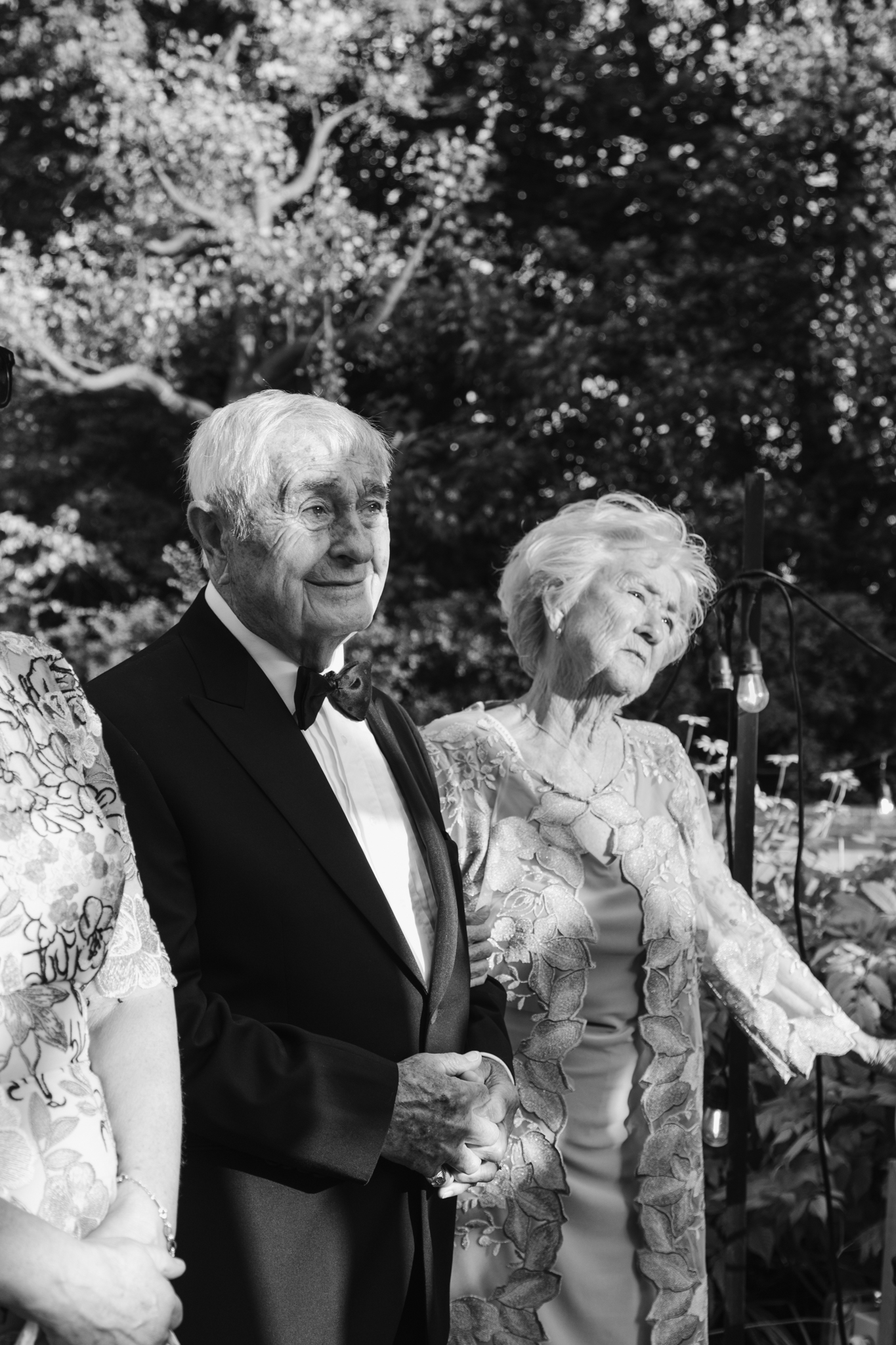 Elderly couple holding hands during a wedding ceremony, captured in black and white in a quiet, photojournalistic moment of emotion and connection