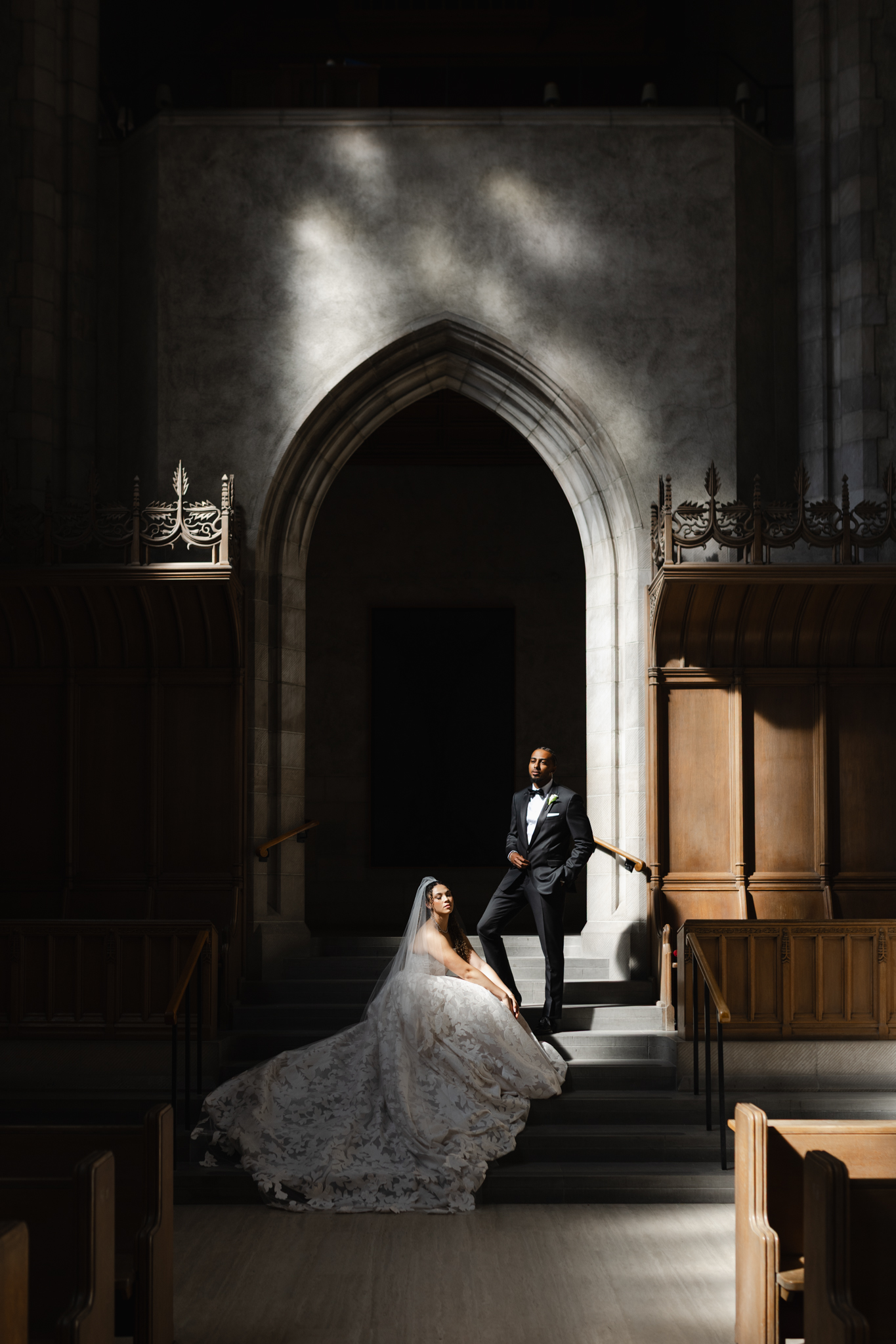 Bride seated on cathedral steps with groom standing above her under dramatic architectural arches, captured in an editorial wedding photography style with sculpted light and refined composition