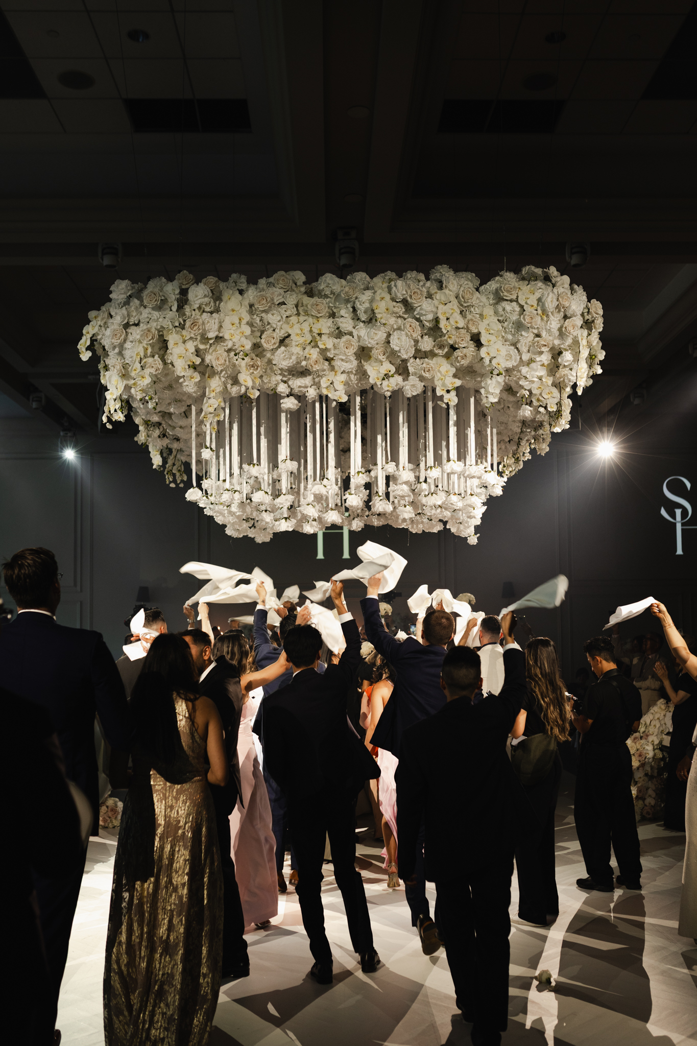 Wedding guests waving napkins in the air during a lively reception under a large floral chandelier, captured in a dynamic, photojournalistic style at Arlington Estate