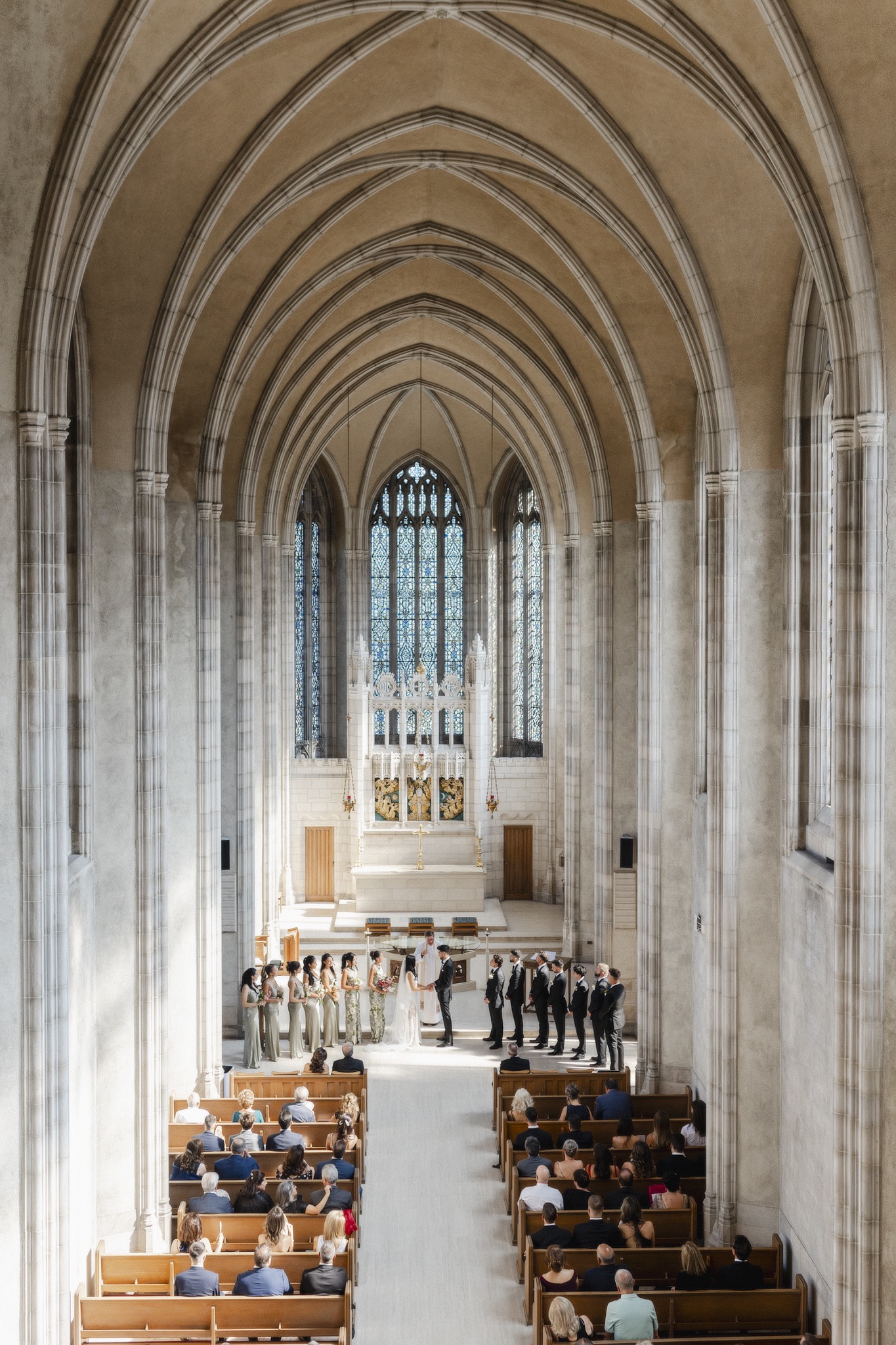 Wide view of a wedding ceremony inside a grand cathedral Trinity College Chapel with soaring arches and guests seated in pews, captured in a timeless, architectural, photojournalistic style.