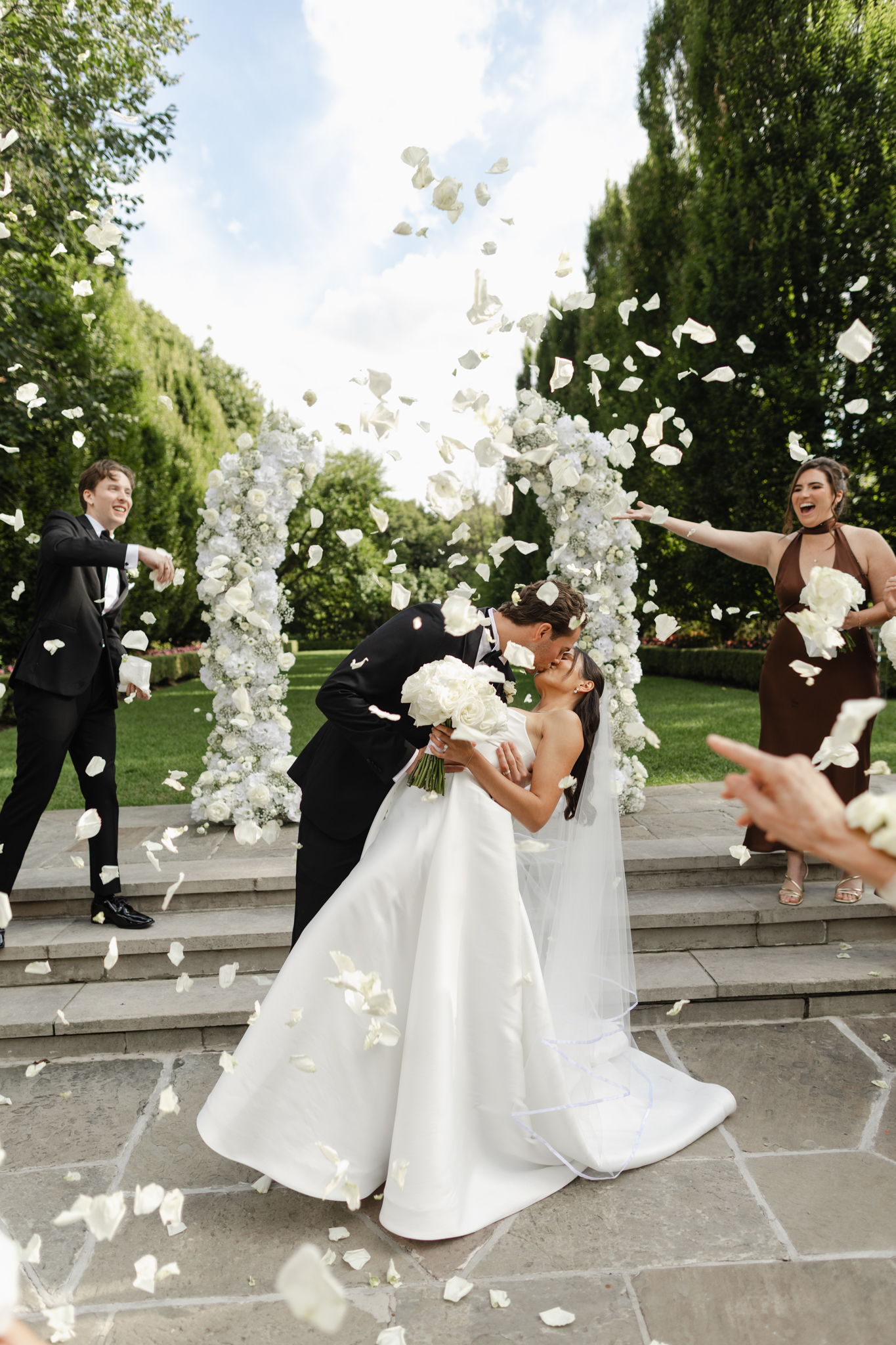 Bride and groom kissing as white flower petals are thrown in the air during an outdoor wedding celebration, captured in a joyful, candid, photojournalistic style in a garden setting.